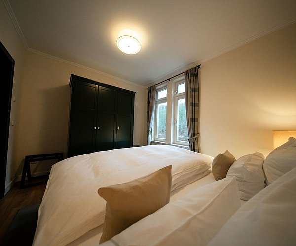 Im Vordergrund ein großes Doppelbett mit vielen Kissen, einem TV an der Wand, mit gemütlichem Licht und großem Fenster mit Blick in den Harz.